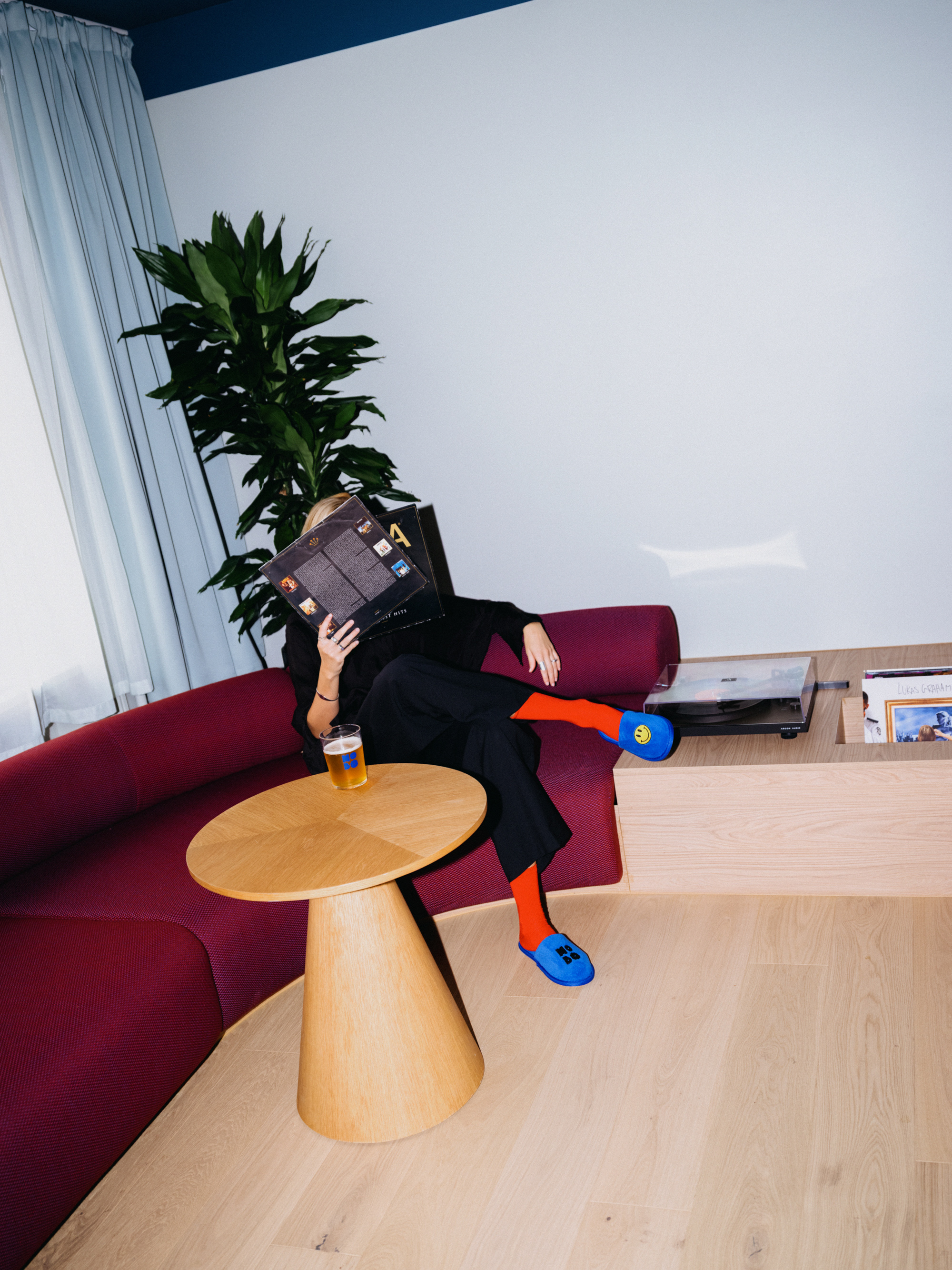 Guest relaxing on a red velvet sofa with a record sleeve in front of the face, in orange socks and blue Hobo slippers