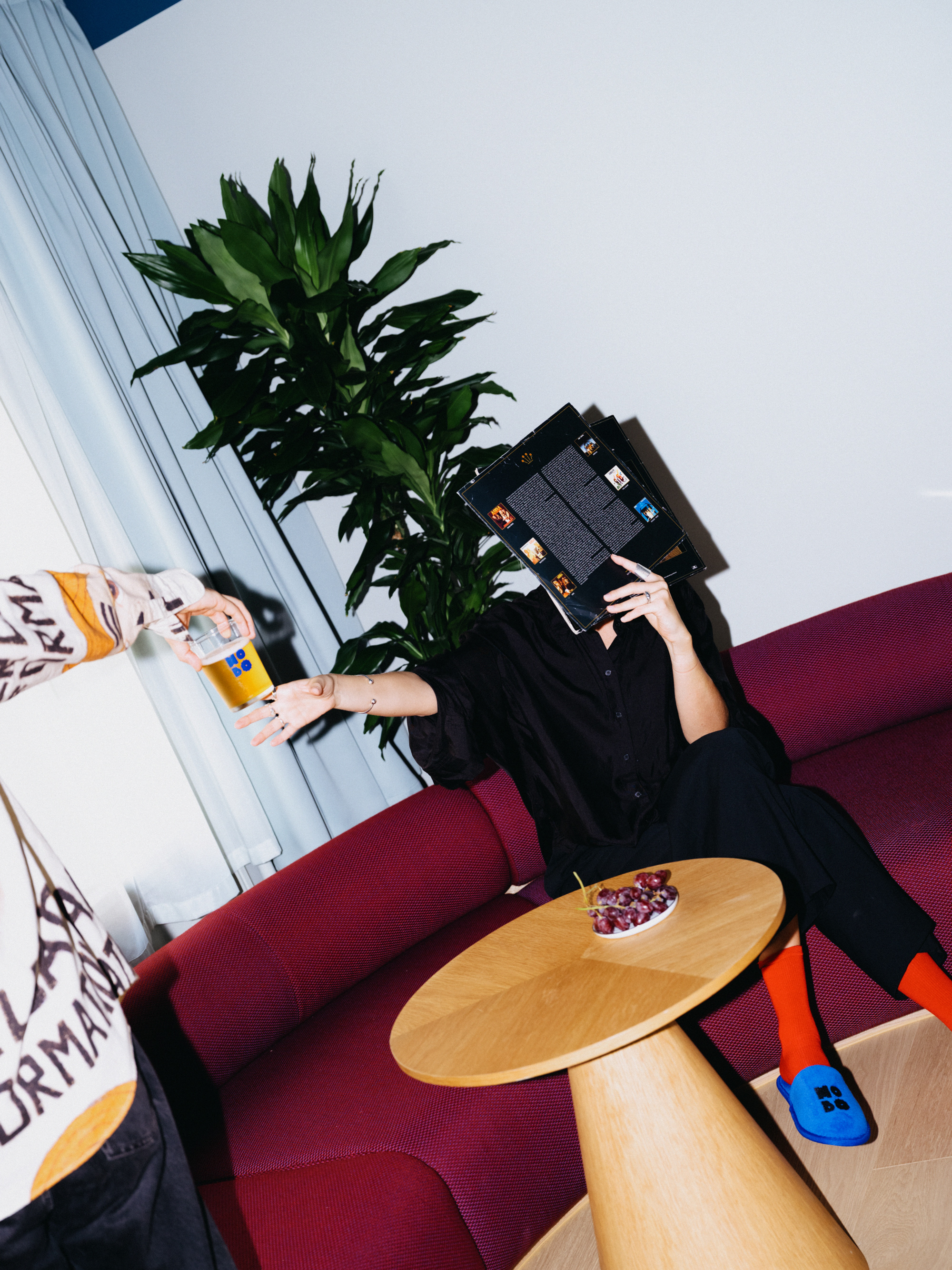 Guest on a red sofa holding up a record sleeve while a friend passes a beer