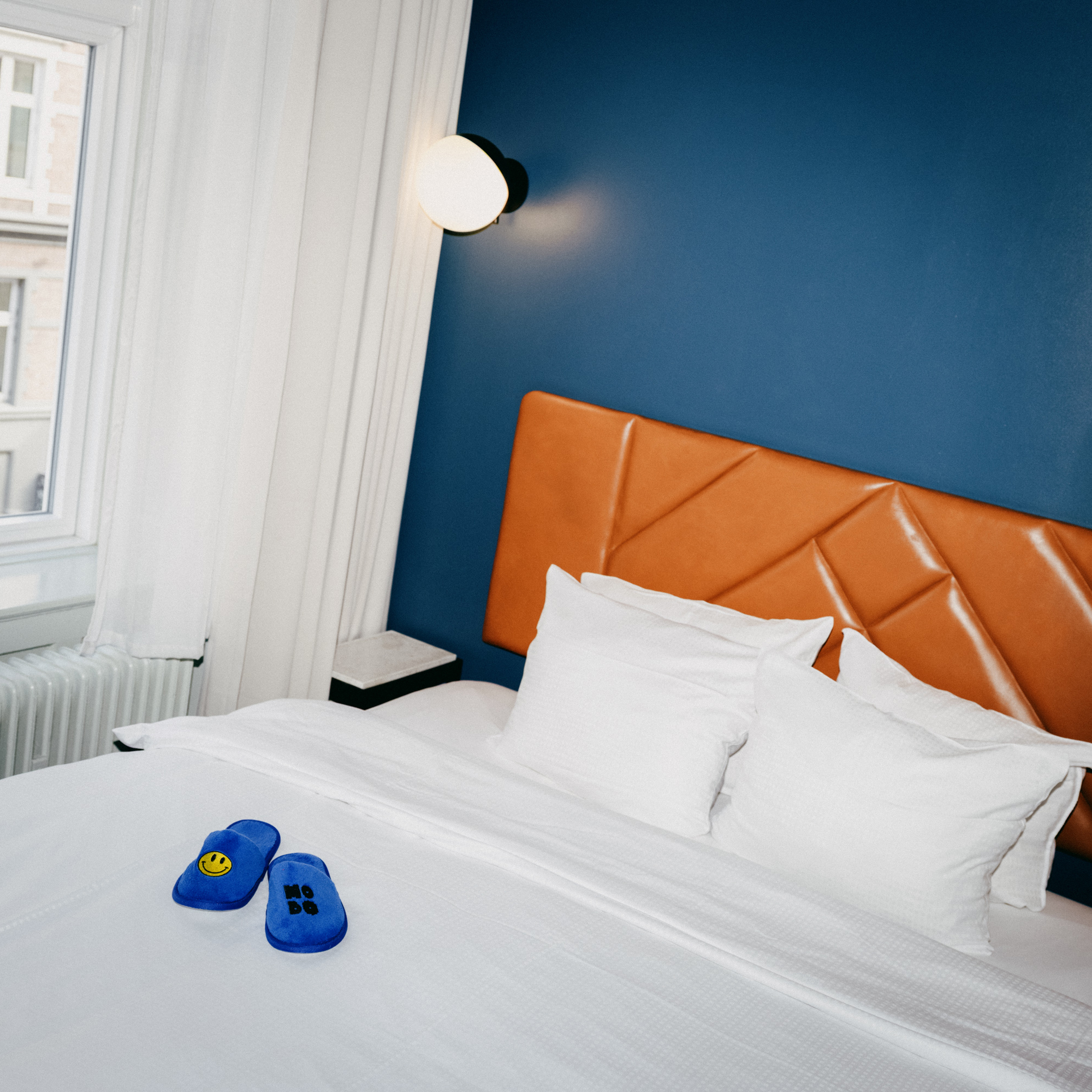 Hotel bed with white linen, a leather headboard and blue Hobo slippers