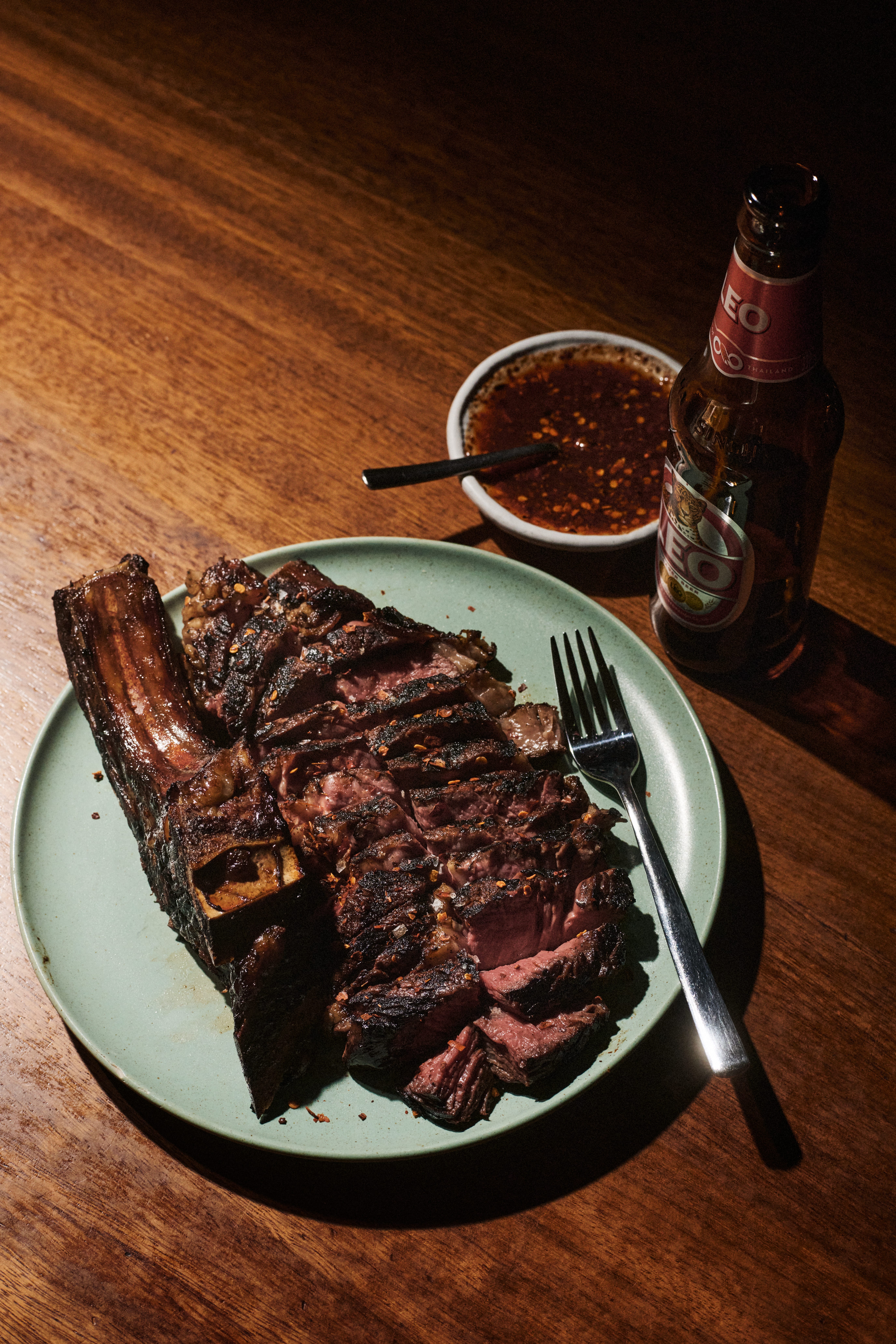 Grillat högrevsben skivat på ett grönt fat, med chilisås och en flaska Leo-öl